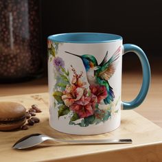 a coffee mug with a hummingbird and flowers painted on it next to a spoon