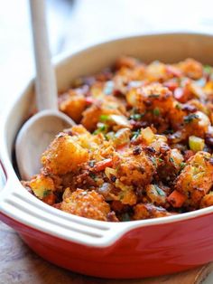 a red casserole dish filled with stuffing and vegetables on a wooden board next to a spoon