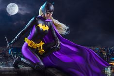 a woman dressed as batman sitting on top of a building at night with the moon in the background