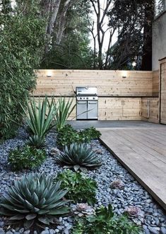 an outdoor kitchen is surrounded by plants and rocks in the foreground, along with a wooden deck