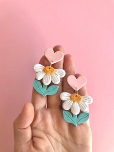 a hand holding two flower shaped earrings on top of a pink surface with hearts in the background