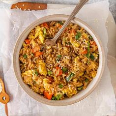 a bowl filled with rice and vegetables on top of paper next to a wooden spoon