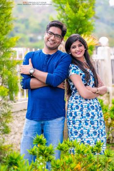 a man and woman standing next to each other in front of some trees with their arms crossed