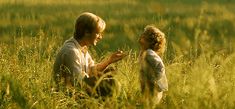 two people sitting in tall grass with one pointing at the other's hand and looking into each others eyes