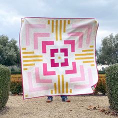 a person holding up a pink and yellow quilt in the middle of some bushes,