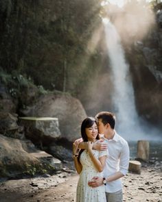 a man and woman standing in front of a waterfall with the sun shining down on them