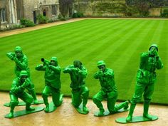 green army figurines are posed in front of a grassy area with an old building in the background