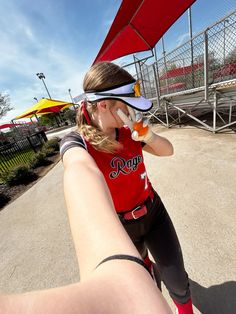 a woman in red shirt and black pants drinking from a bottle while standing next to a fence