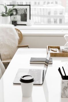 a white table with a laptop, pen and cup on it in front of a window