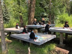 several children are sitting at picnic tables in the woods with chalk drawings on them and surrounded by trees