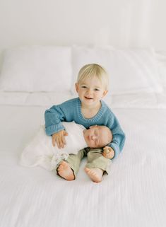 a little boy sitting on top of a bed next to his baby sister, who is holding him