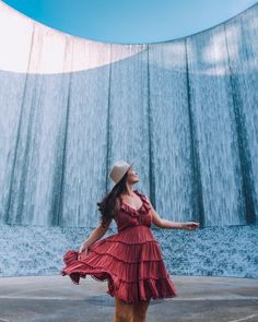 a woman standing in front of a waterfall wearing a red dress and hat with her arms outstretched