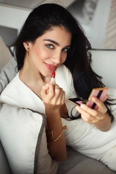 a woman sitting on a couch holding a cell phone and looking at her lipstick in the mirror