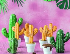 some fake cactus plants in small white pots on a pink surface with palm leaves and a purple wall behind them