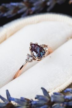 a close up of a ring in a box with lavender flowers around it and an oval diamond on the side