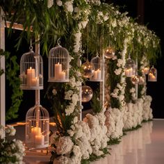 a row of birdcages filled with white flowers and candles next to greenery