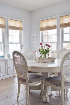 a dining room table with four chairs and a basket on top of it, in front of two windows