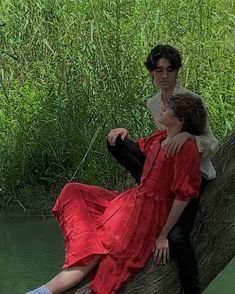 two people are sitting on a tree branch in the water and one person is wearing a red dress