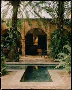 a pool surrounded by palm trees and greenery in front of a brick building with arched doorways