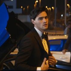 a man in a tuxedo standing next to a car at night with other people