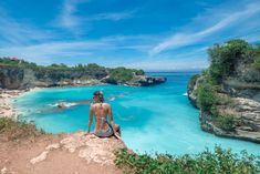 a woman sitting on top of a cliff next to the ocean with clear blue water