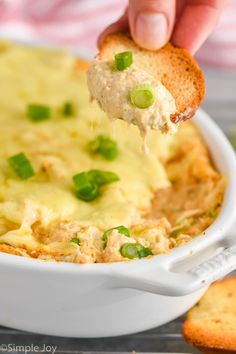 a hand holding a cracker over a casserole dish with chicken and cheese