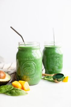 two mason jars filled with green smoothie next to sliced avocado and mango