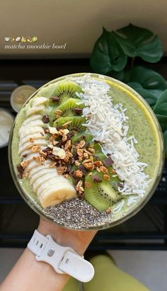 a person holding up a green smoothie topped with bananas, kiwi and nuts