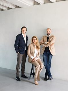 three people standing next to each other in front of a white wall and one person sitting on a chair