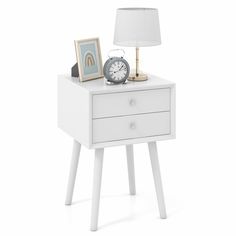 a white night stand with a clock, lamp and pictures on the side table next to it