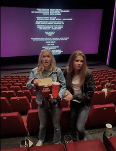 two women sitting on red seats in front of a projection screen holding popcorn buckets