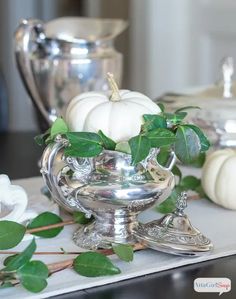 white pumpkins and greenery sit in a silver bowl on a table with other dishes