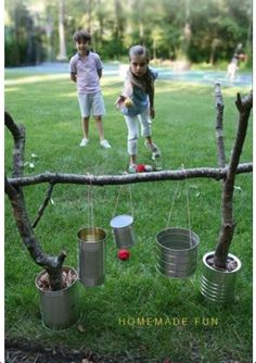 two children playing with tin cans on a tree branch in the grass, and one child is holding an apple