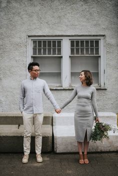 a man and woman holding hands standing in front of a white building with two windows