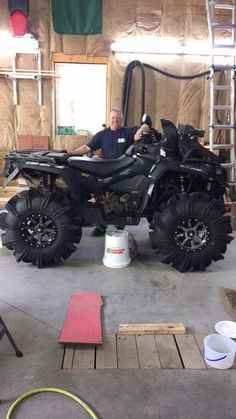 a man is sitting on an atv in a building with large wheels and tires, while another man stands next to the vehicle