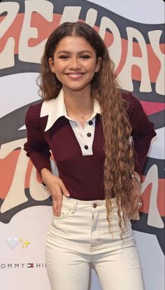 a woman with long hair standing in front of a wall wearing white pants and a maroon shirt