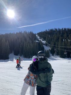 two snowboarders embracing each other in the middle of a snowy area with pine trees behind them