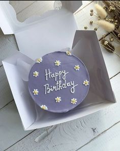 a purple birthday cake with daisies on it in a white gift box next to dried flowers