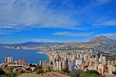 the city is next to the ocean and mountains in the distance, with blue skies above it