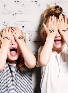 two young children covering their eyes with their hands while standing next to each other in front of a white brick wall