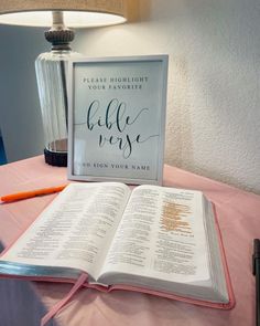 an open bible sitting on top of a table next to a lamp and a pen