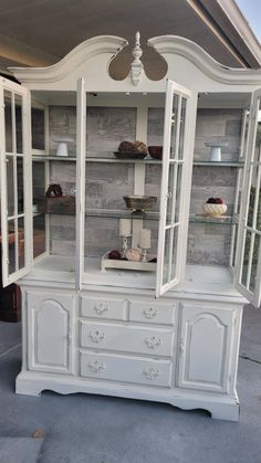 a white china cabinet with glass doors and shelves on the top, in front of a house