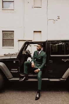 a man in a green suit sitting on the back of a black jeep parked next to a white building