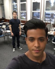 two men standing in a classroom with desks, chairs and one is looking at the camera