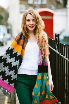 a woman wearing green pants and a white top is holding a colorful knitted shawl