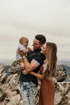 a man and woman holding a baby while standing on top of a rocky hill with mountains in the background