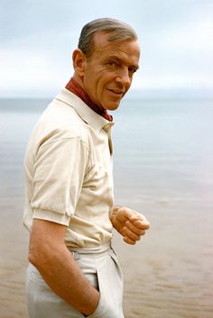 an older man standing on the beach with his hands in his pockets and looking at the camera