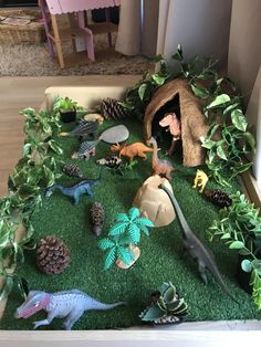 an assortment of toy dinosaurs in a play area with fake plants and pine cones on the floor