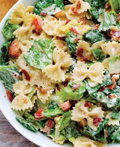 a white bowl filled with pasta salad next to tomatoes and lettuce on a wooden table