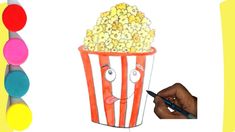 a hand holding a marker next to a drawing of a popcorn bucket with eyes drawn on it